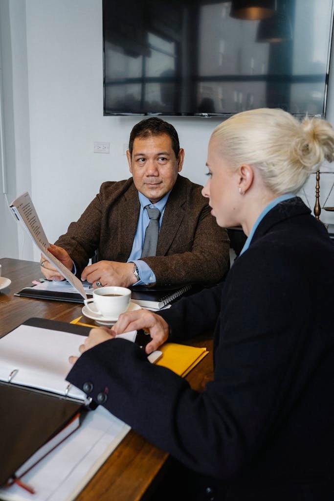 Side view of serious woman in formal clothes sitting at table and discussing details of project with ethnic colleague