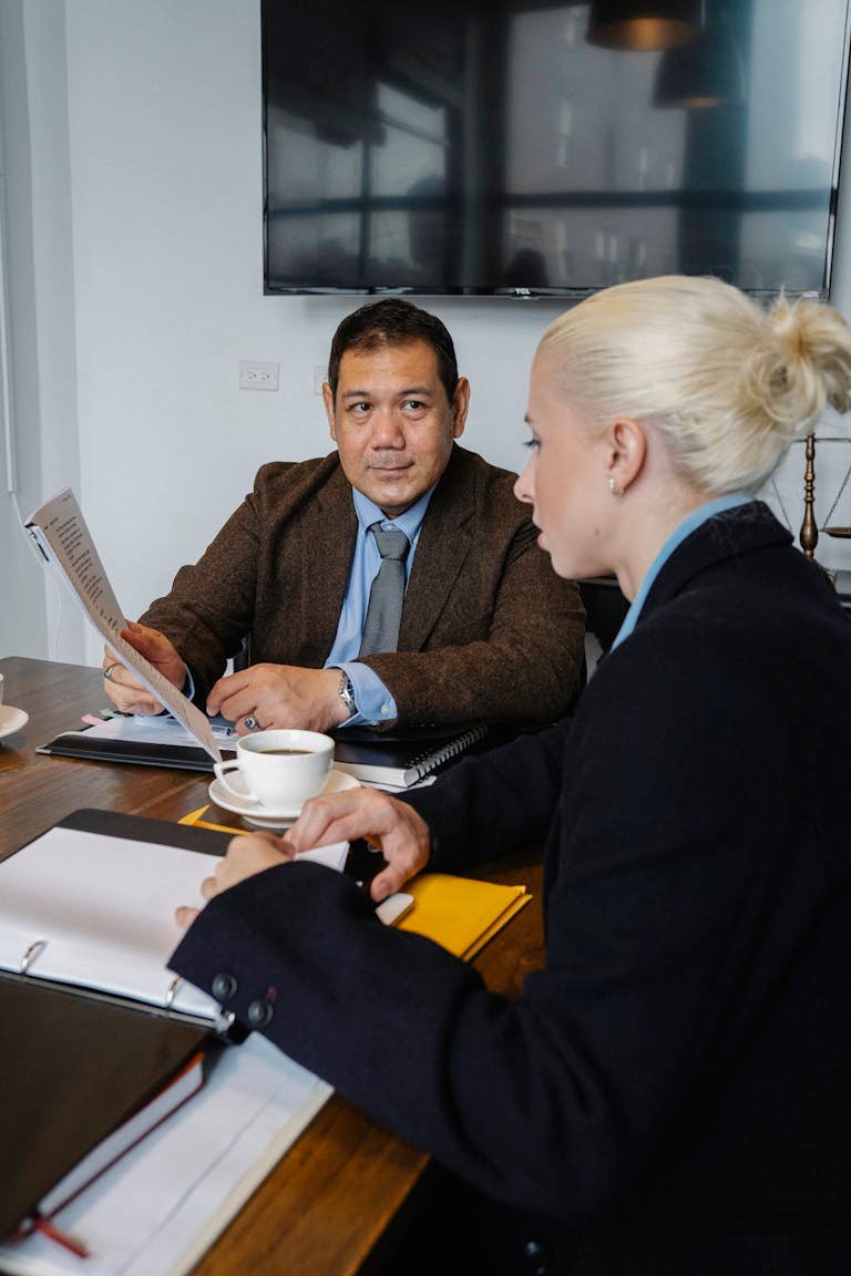 Side view of serious woman in formal clothes sitting at table and discussing details of project with ethnic colleague