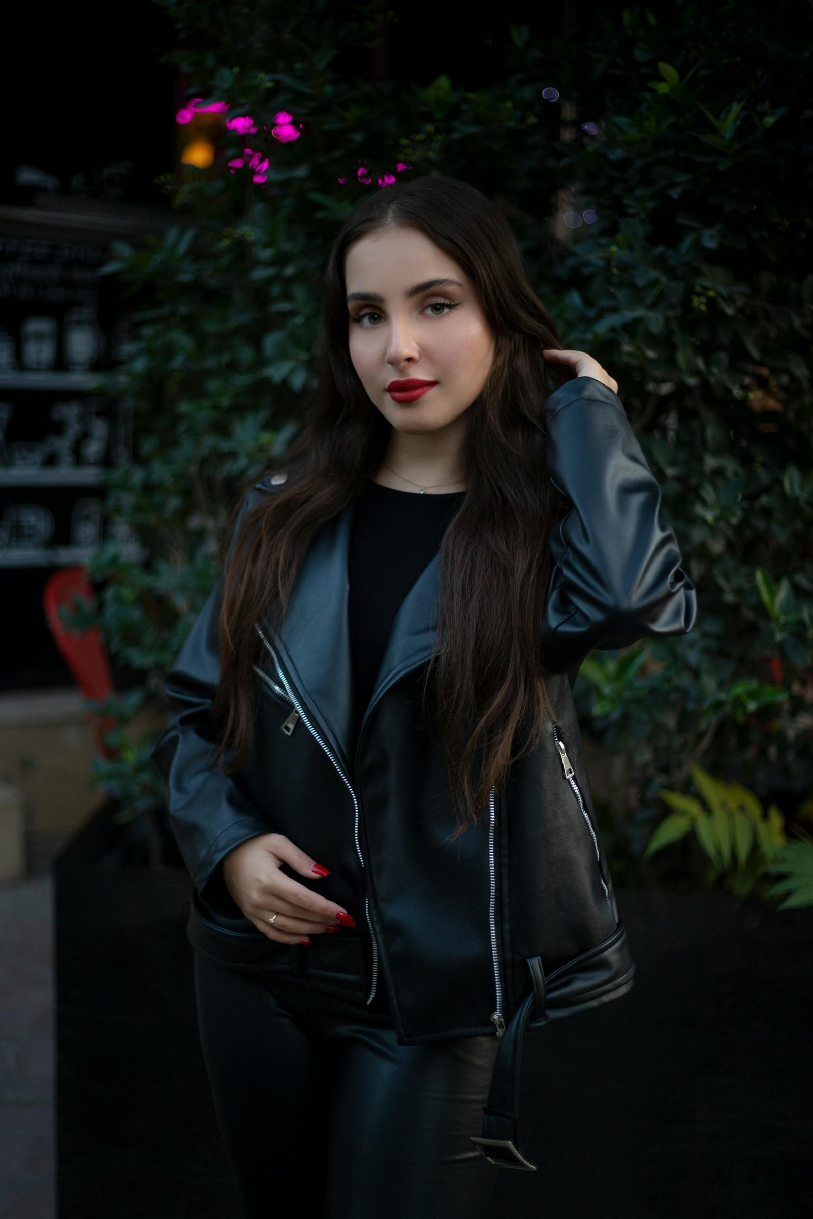 Stylish woman with red nails and leather jacket posing outdoors at night.